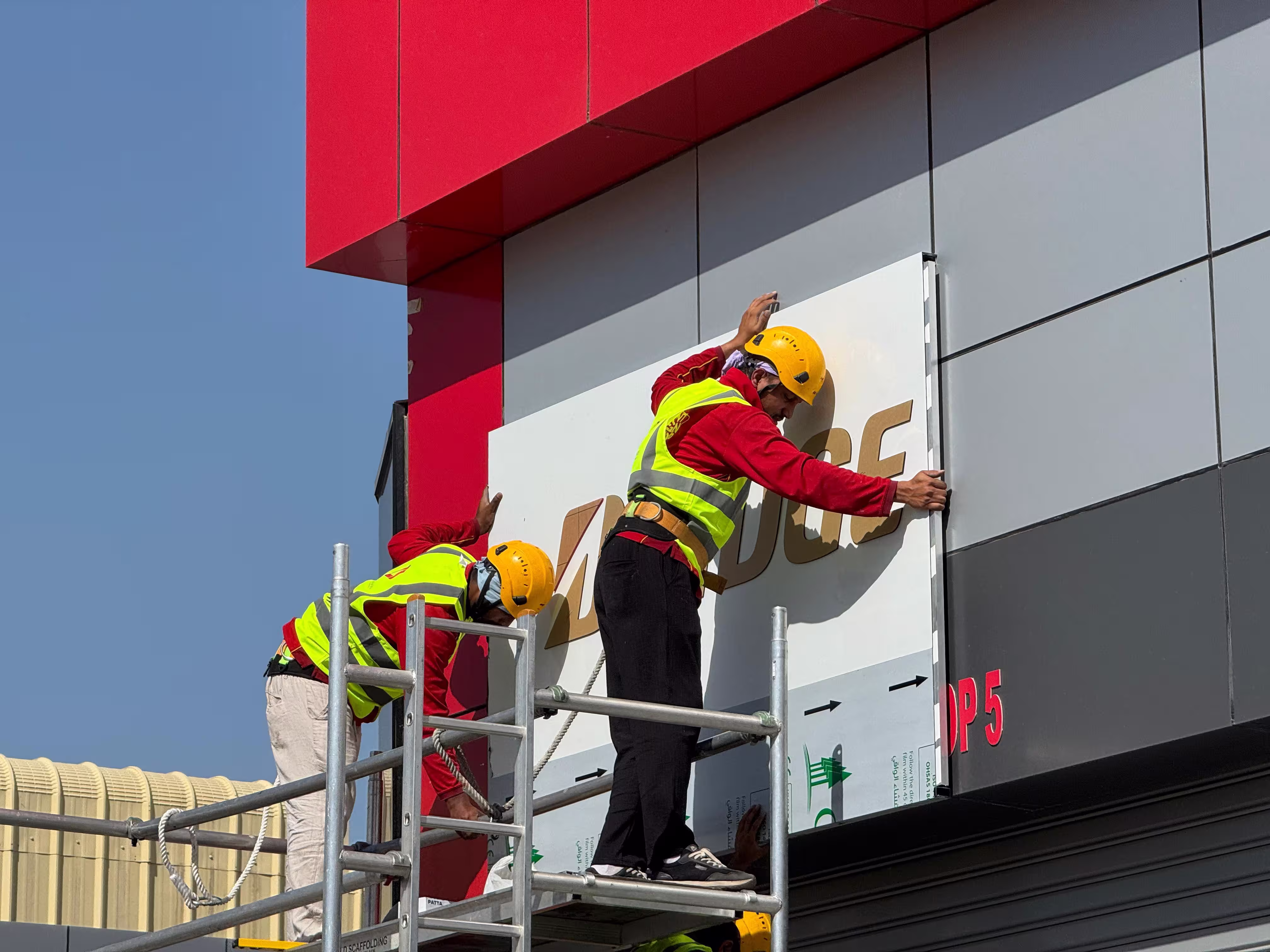 Two workers in safety gear installing a large exterior signage panel on a modern building facade